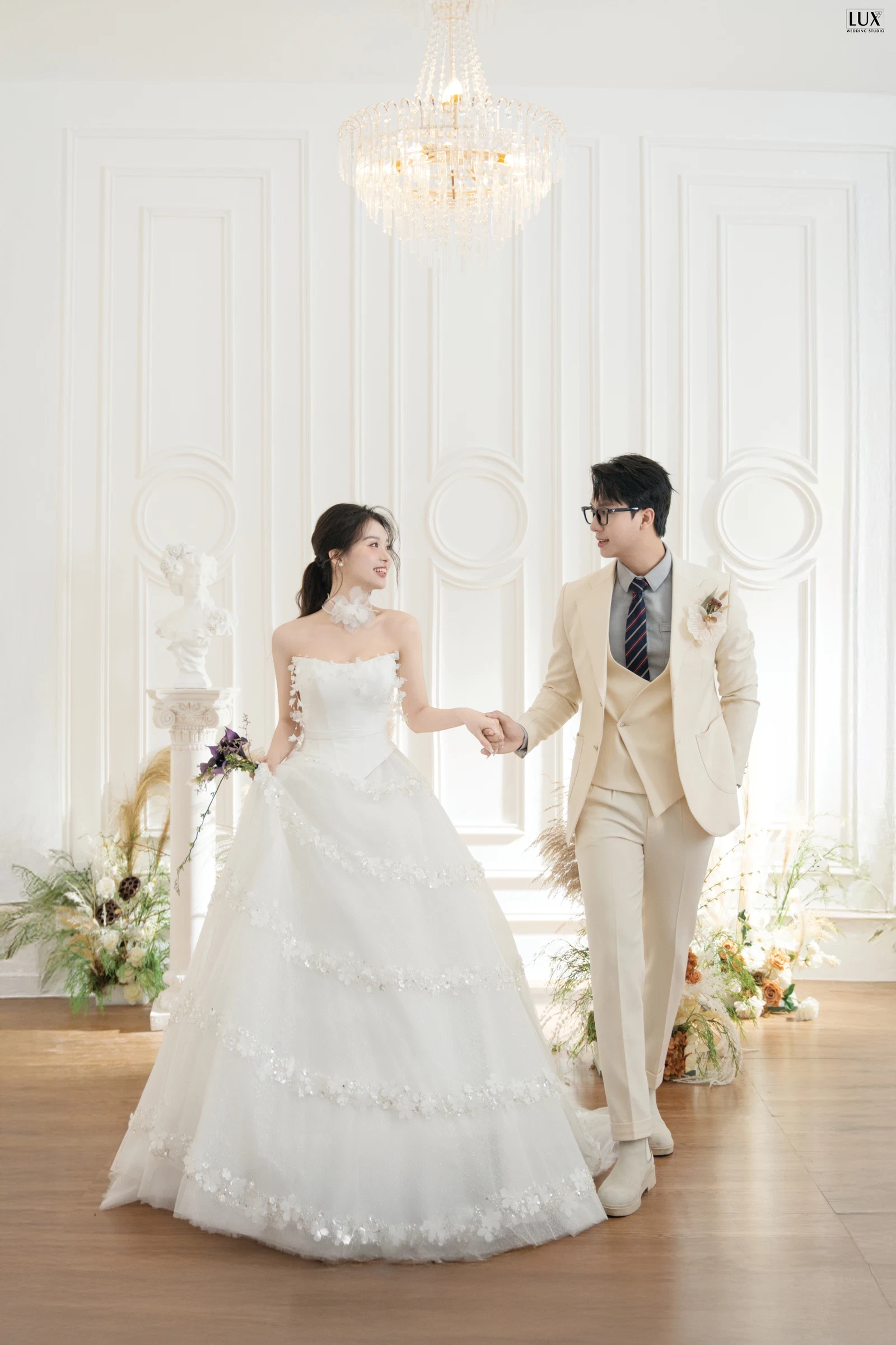 Cô dâu váy cưới trắng bồng xòe nắm tay chú rể vest màu kem bước đi trong phòng tường phào trắng có đèn chùm pha lê và hoa khô hai bên
