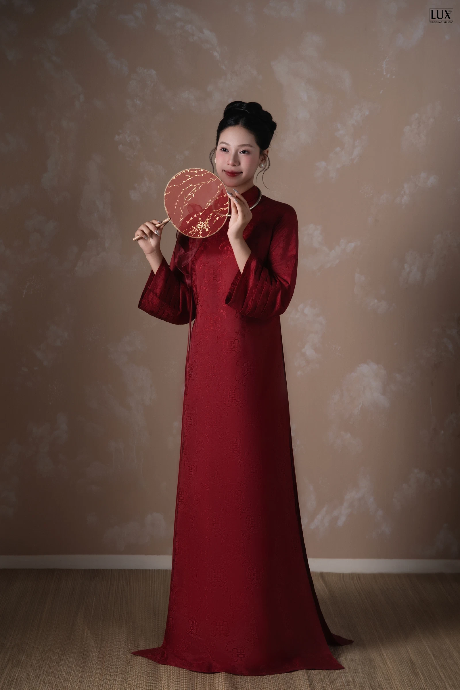 Cô dâu áo dài đỏ trơn cổ cao, tóc búi gọn, hai tay cầm quạt tròn đỏ thêu hoa, đứng trước phông nâu loang