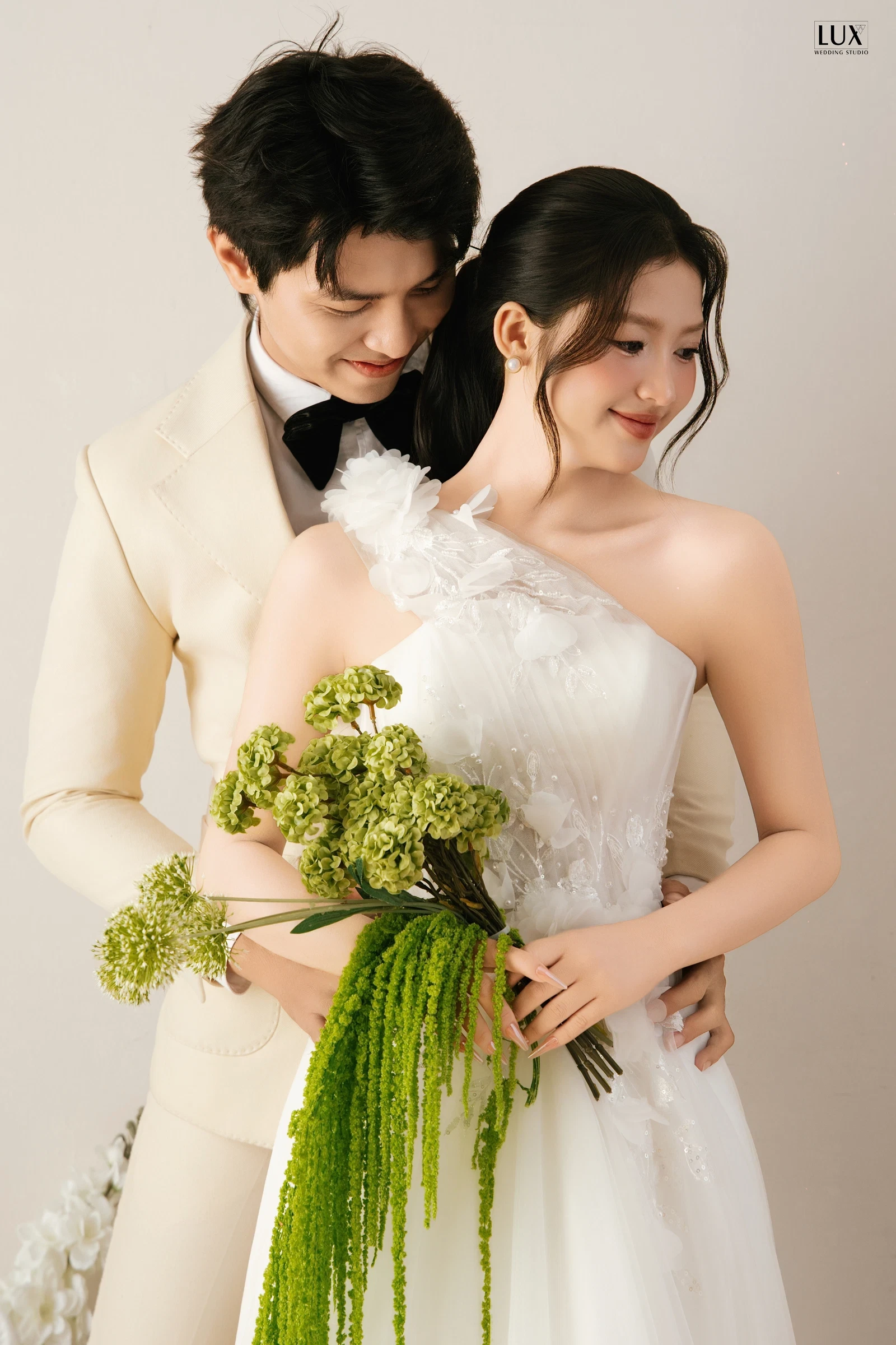 Chú rể vest kem nơ đen ôm cô dâu từ phía sau, cô dâu váy trắng một vai cầm bó hoa xanh rủ dài trên nền be