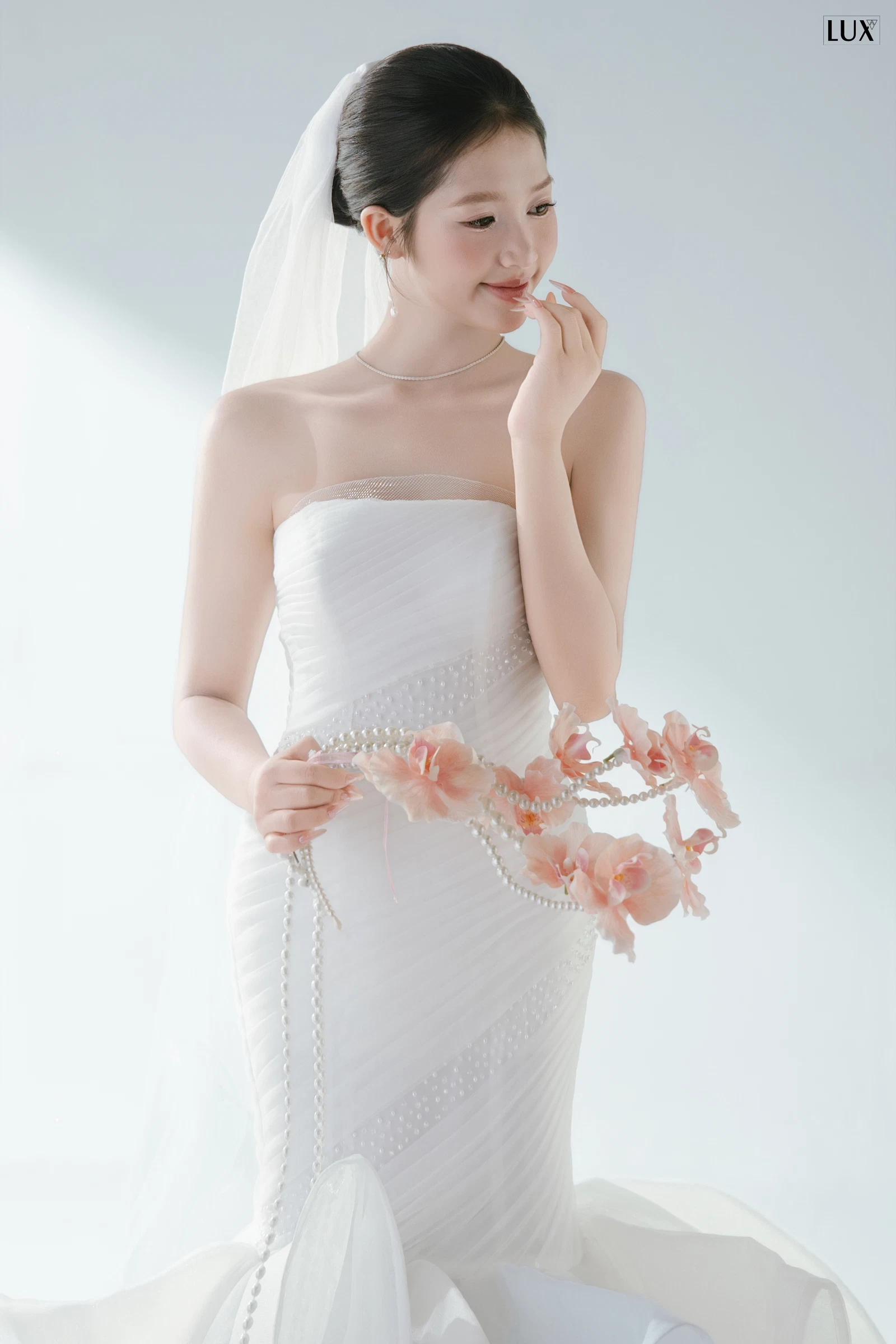 Cô dâu búi tóc thấp cài voan, trang điểm trong trẻo, mỉm cười đưa tay lên môi, mặc váy cúp ngực trắng