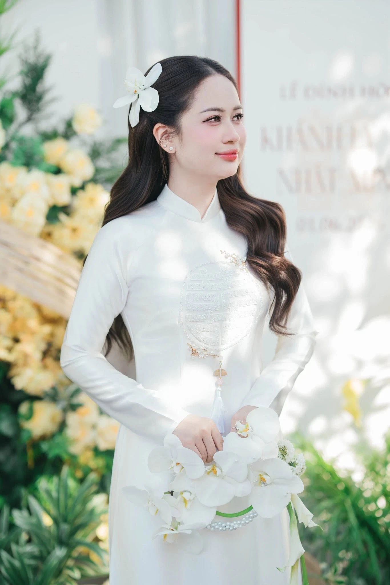 Cô dâu áo dài đỏ cách tân tay loe phủ ren, đội khăn voan đỏ, đứng cạnh ma-nơ-canh mặc áo dài đỏ thêu hoa và bó hoa đỏ cắm trên giá