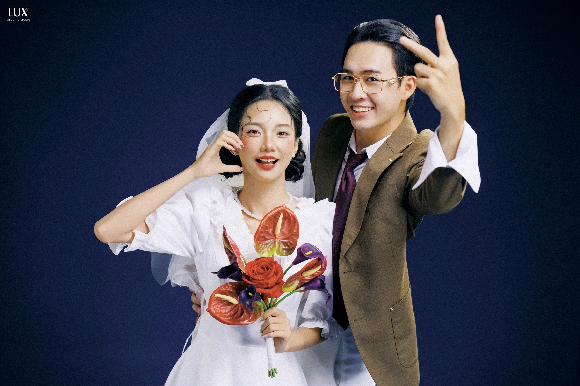 Cô dâu đội voan, váy trắng tay bồng, ôm bó hoa đỏ, cười tinh nghịch cạnh chú rể đeo kính, áo vest nâu trước phông xanh đậm