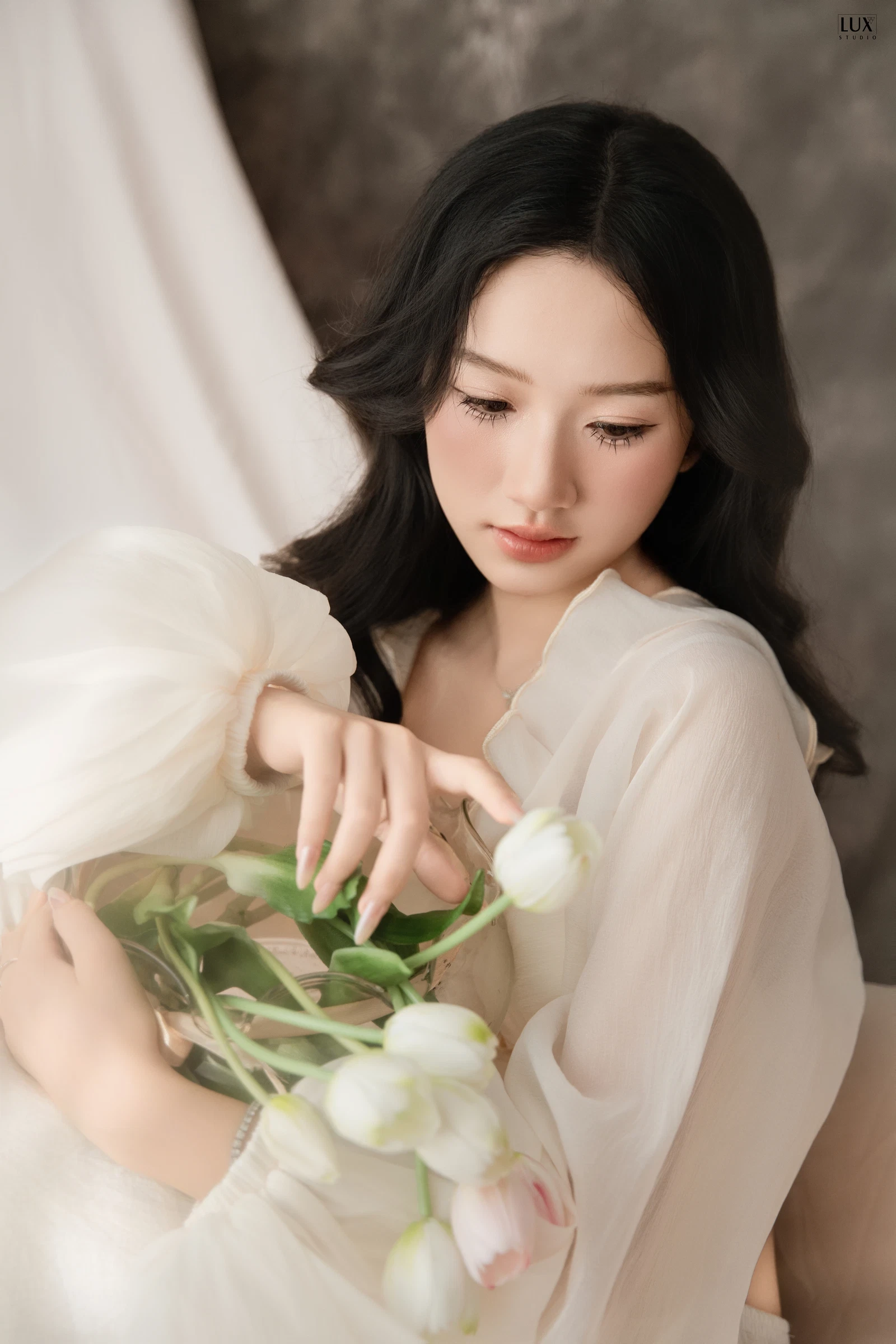 Cô gái tóc đen dài mặc áo voan trắng tay bồng cúi nhìn, tay khẽ chạm vào bó hoa tulip trắng ôm trong lòng