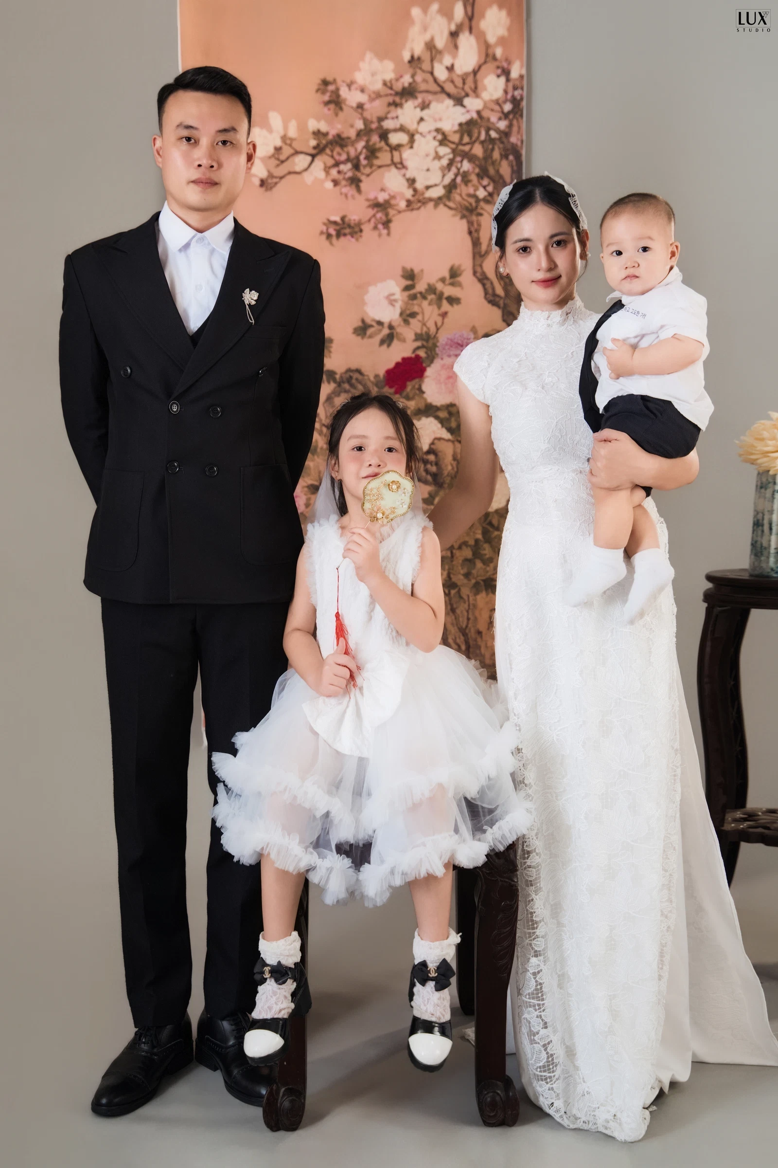 Bố mặc vest đen, bé gái váy trắng xòe bồng cầm quạt nhỏ, mẹ mặc áo dài ren trắng bế bé trai, cả nhà đứng trước tấm tranh hoa đào