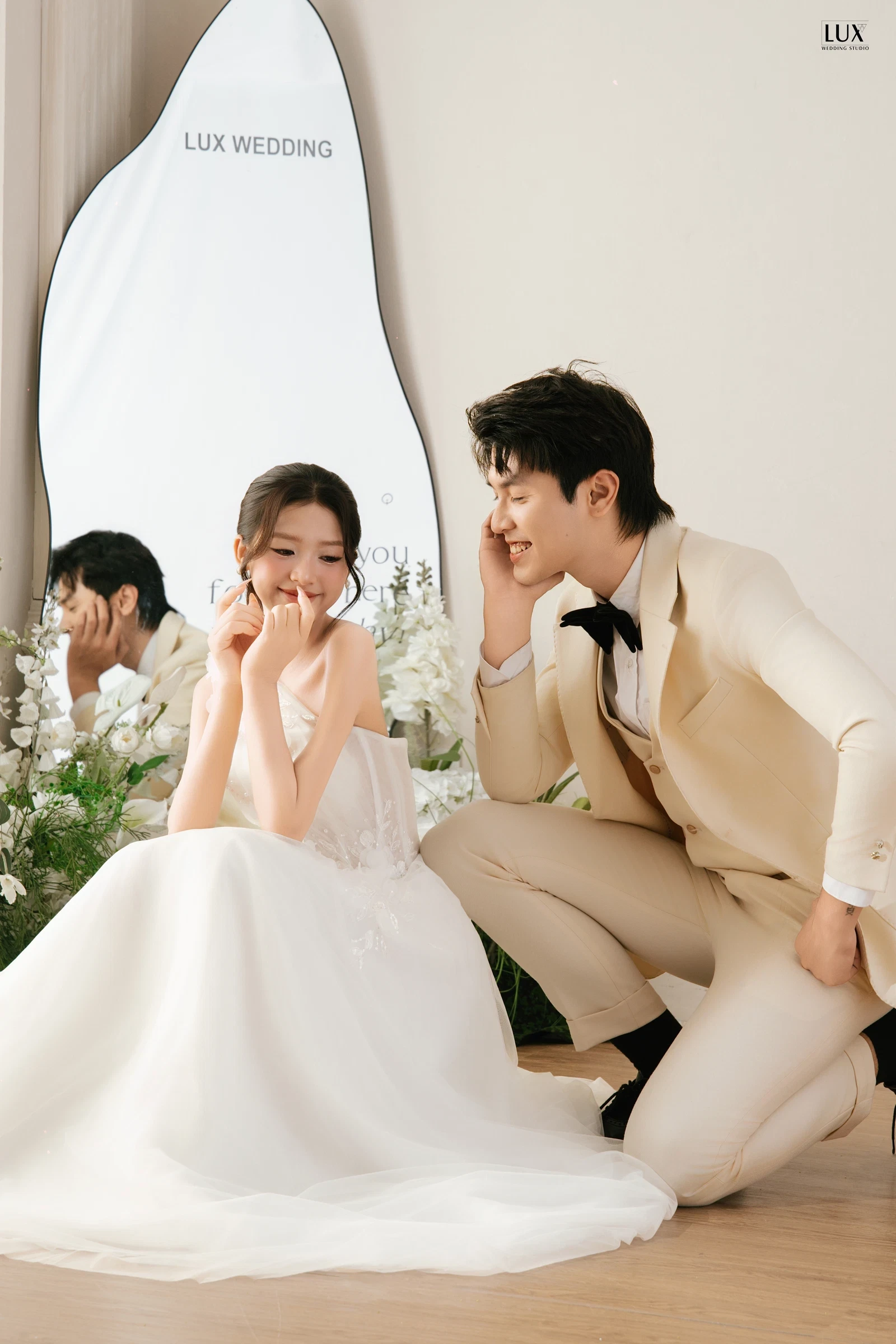 Cô dâu váy trắng ngồi giữa hoa trắng, tay chạm cằm mỉm cười; chú rể vest màu kem nơ đen ngồi cạnh nhìn cô dâu, phía sau là chiếc gương lớn