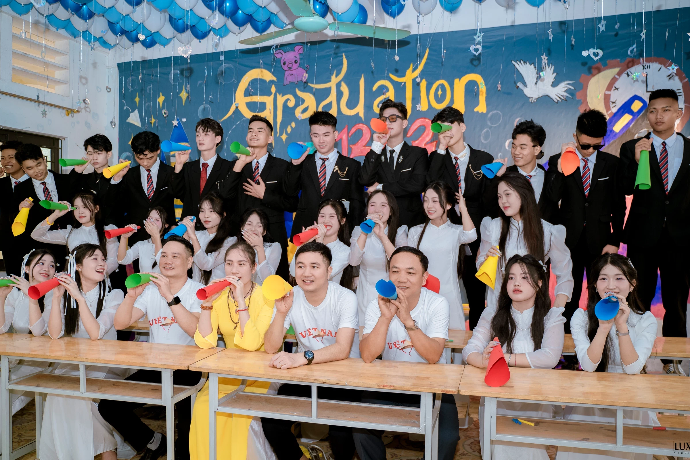 Cả lớp và thầy cô cùng cầm loa giấy nhiều màu tạo dáng vui nhộn trước phông Graduation trong lớp học