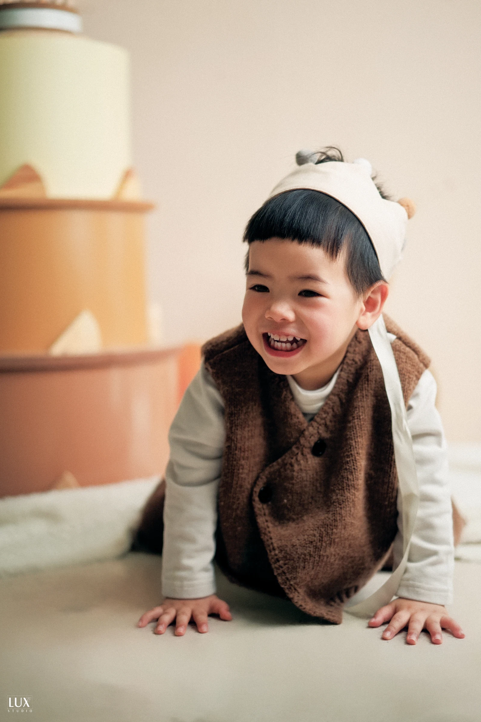 Bé trai đội mũ vải, mặc gile len nâu bò trên sàn và cười tươi, phía sau là mô hình bánh sinh nhật nhiều tầng