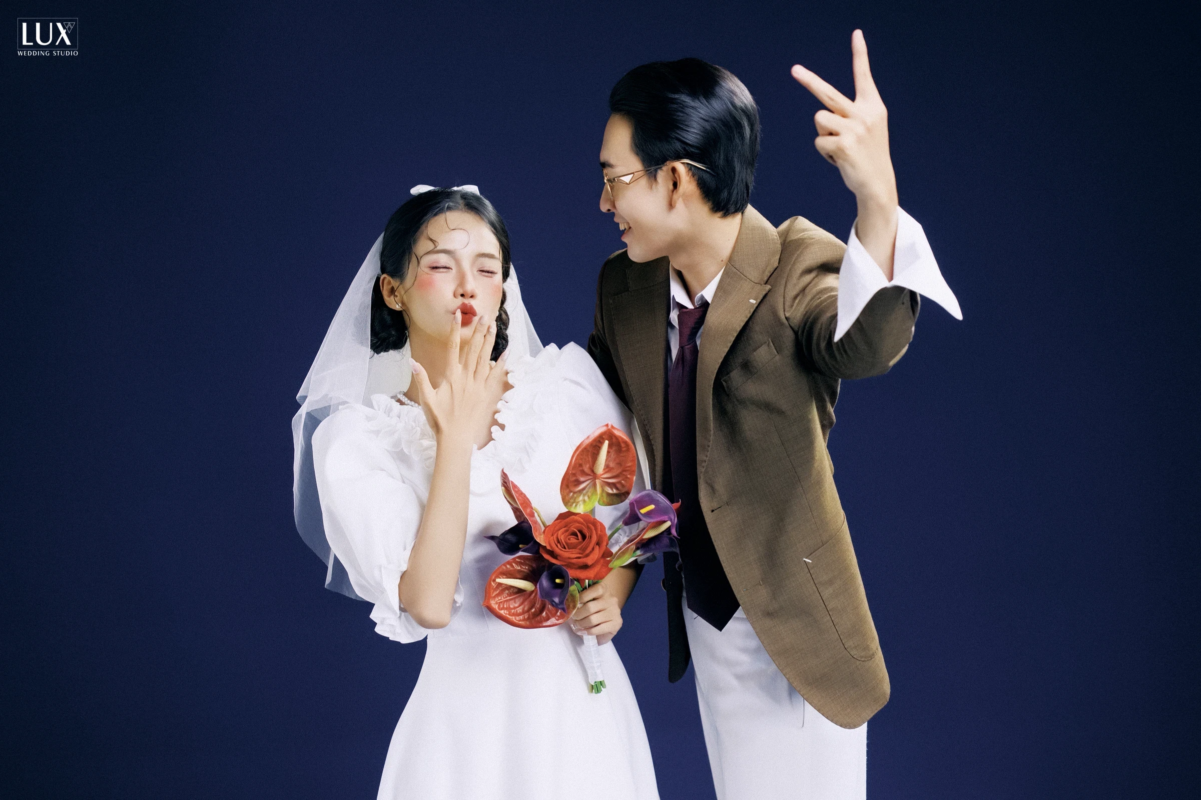 Cô dâu váy trắng mạng ngắn cầm bó hoa đỏ, chú rể kính tròn vest nâu giơ tay tạo dáng vui trên phông xanh navy