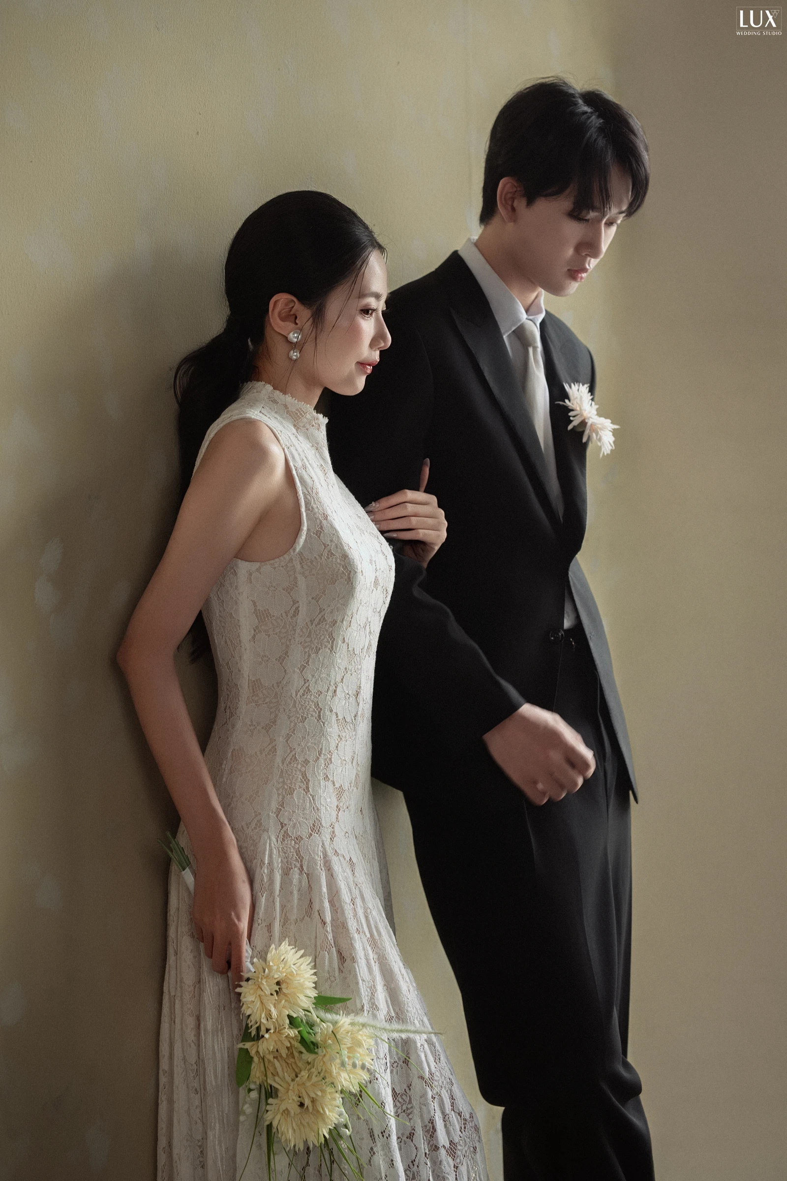 Chú rể vest đen đứng cạnh cô dâu váy ren trước phông be