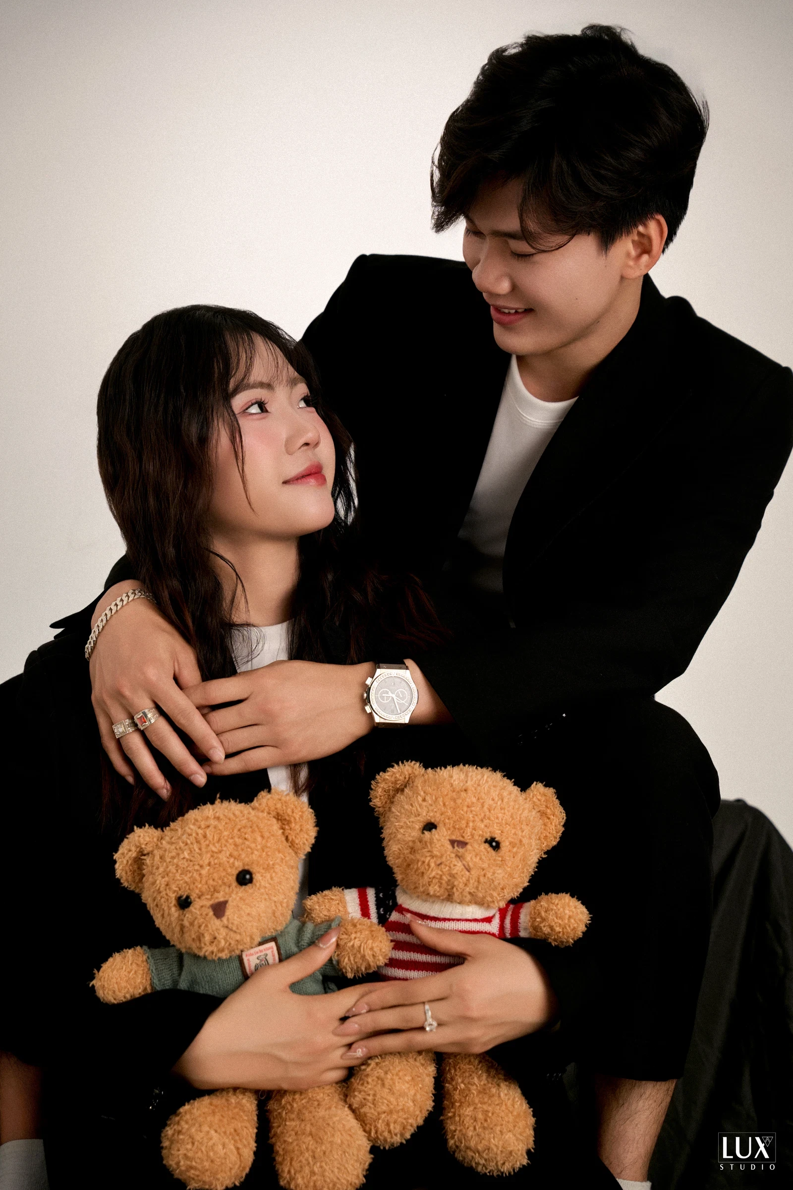 Chàng trai mặc vest đen ôm cô gái từ phía sau, cô gái ôm hai chú gấu bông, cả hai nhìn nhau cười trước phông xám