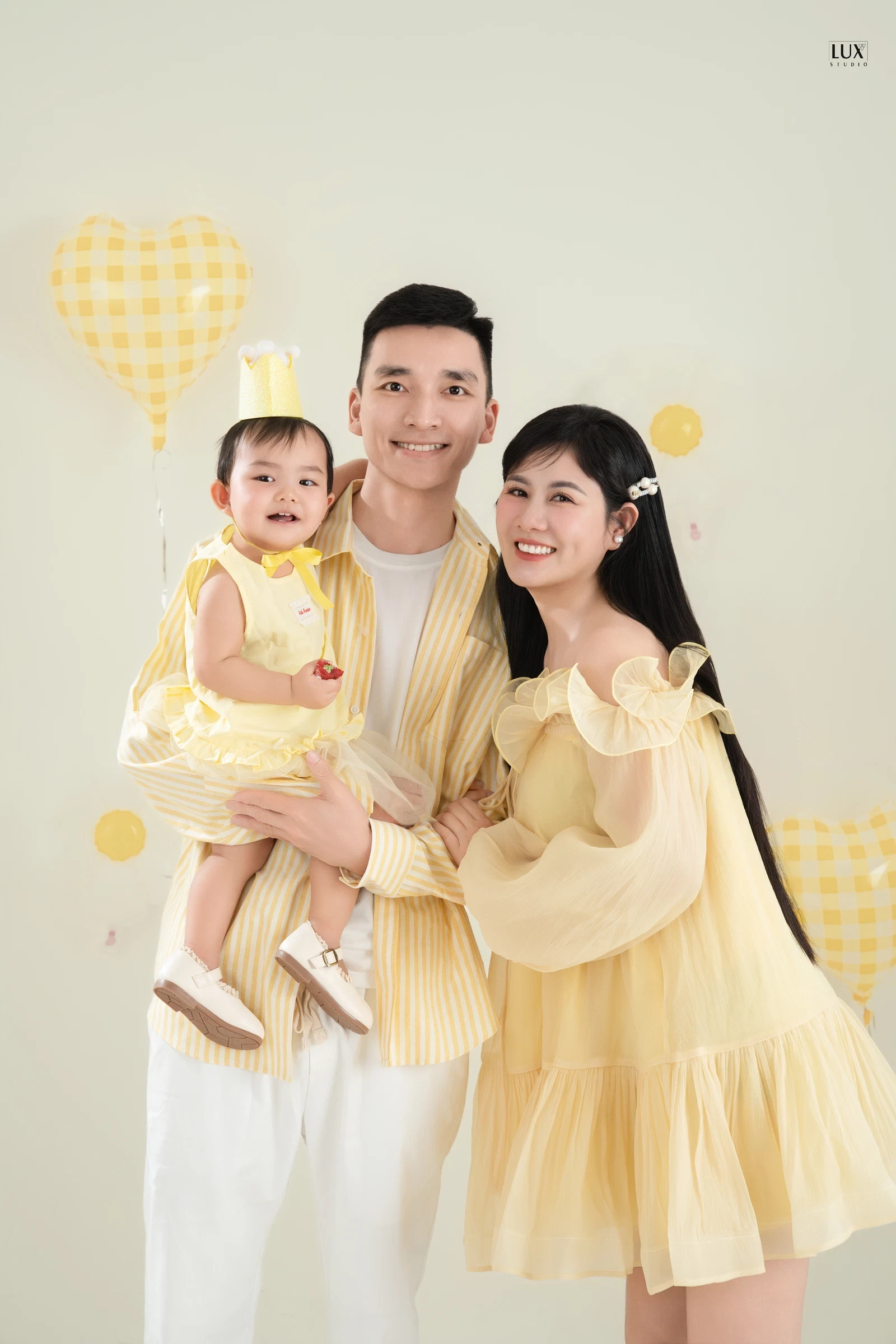 Bố bế bé gái đội vương miện giấy, mặc đồ vàng nhạt, mẹ váy vàng trễ vai tựa vào bố, cả nhà cười trước phông kem có bóng bay hình trái tim