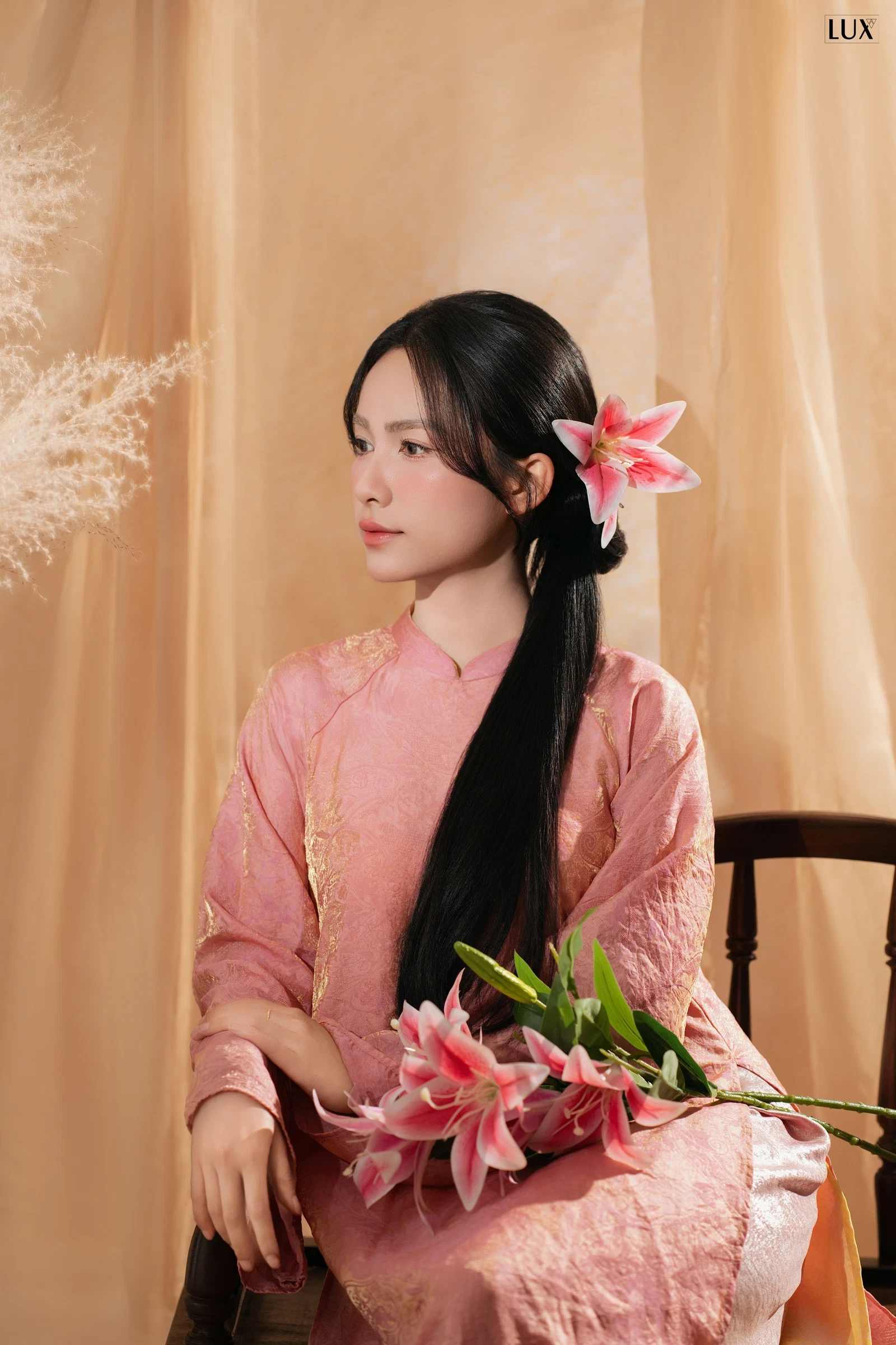 Cô gái mặc áo dài hồng, tóc dài cài hoa ly, ngồi ghế gỗ cầm bó hoa ly hồng trước phông rèm màu be