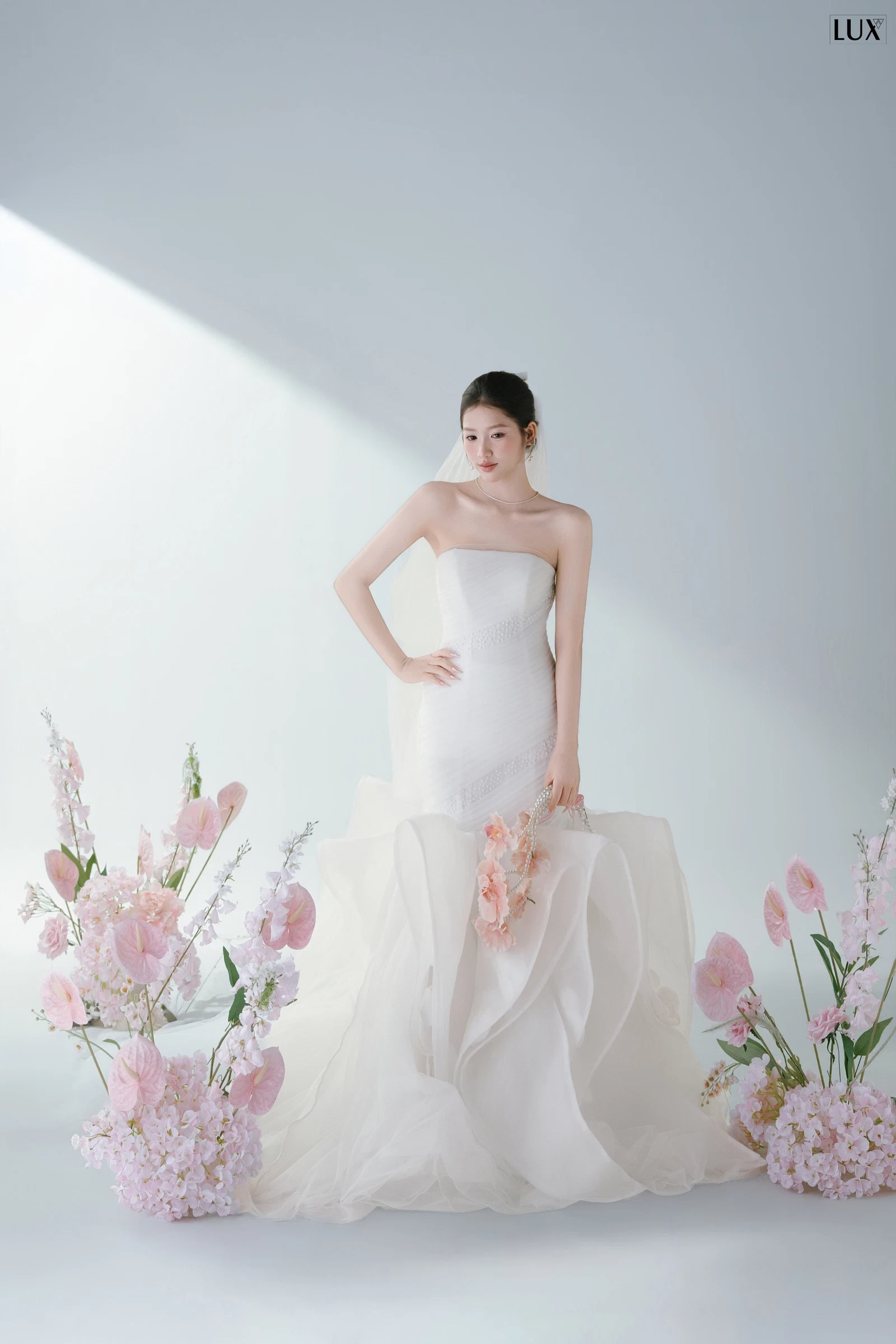 Cô dâu váy cưới quây ngực đuôi xếp tầng đứng giữa nền trắng vô cực, hai bên là những cụm hoa hồng phấn