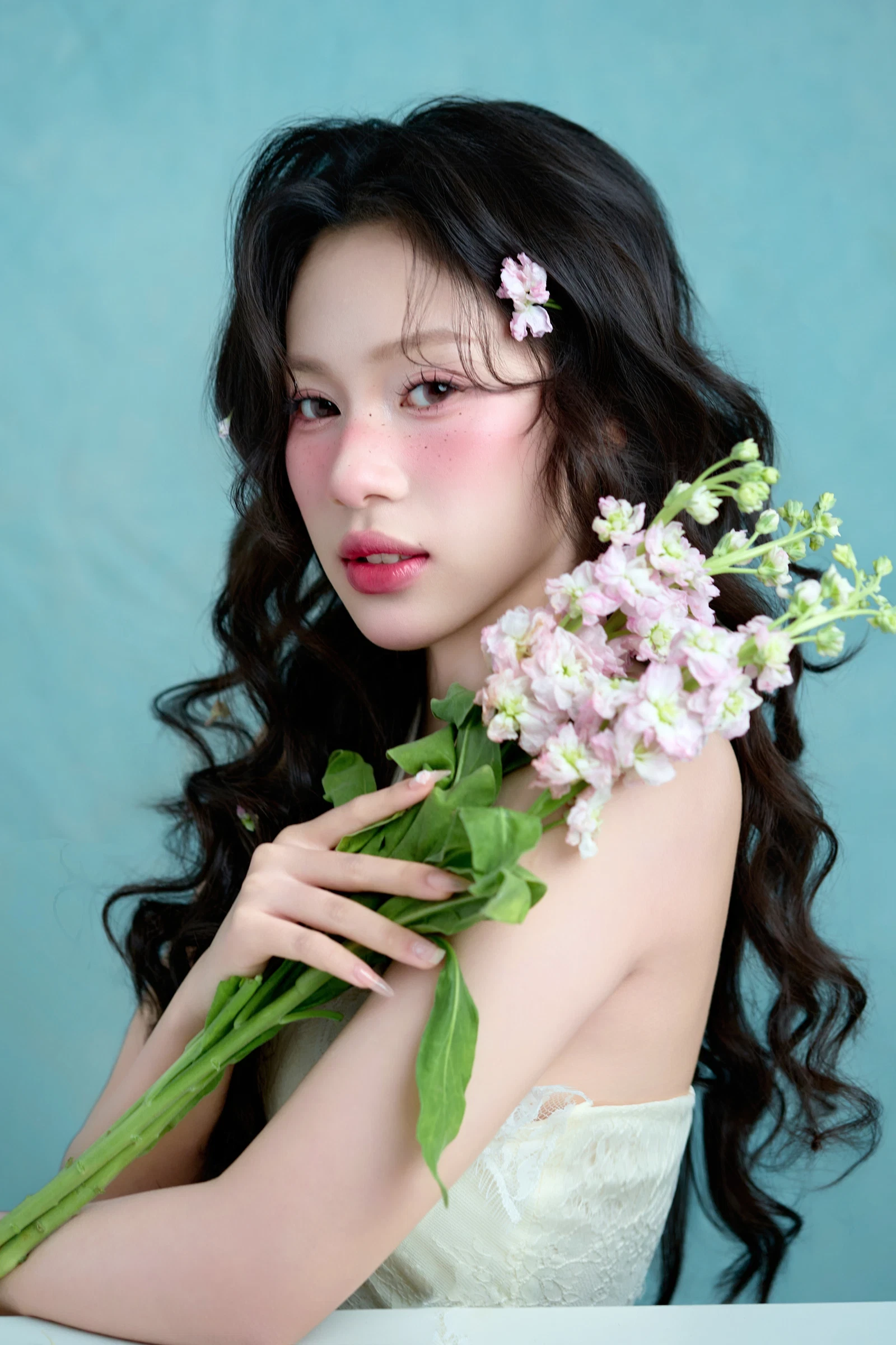 Cô gái tóc đen xoăn dài cài hoa nhỏ, má hồng, ôm bó hoa tím nhạt và nhìn thẳng vào ống kính trước phông nền xanh