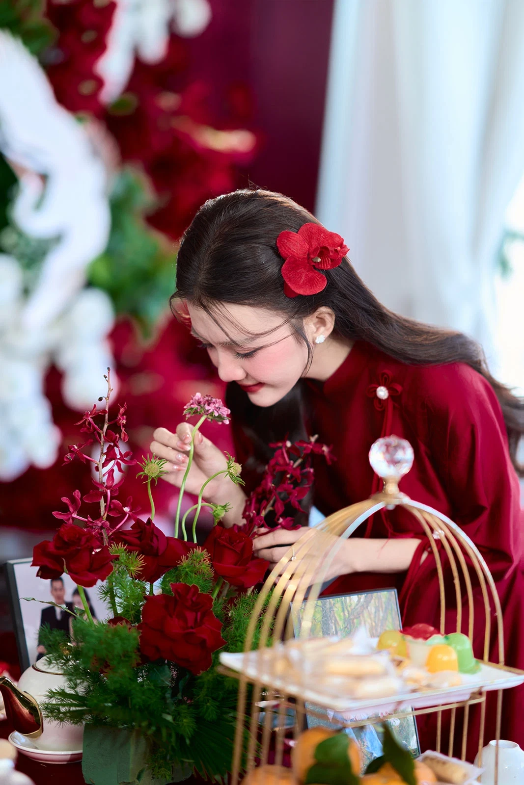 Cô dâu áo dài đỏ cài hoa lan đỏ trên tóc, cúi xuống chỉnh lọ hoa hồng đỏ bên khay bánh trong phòng lễ