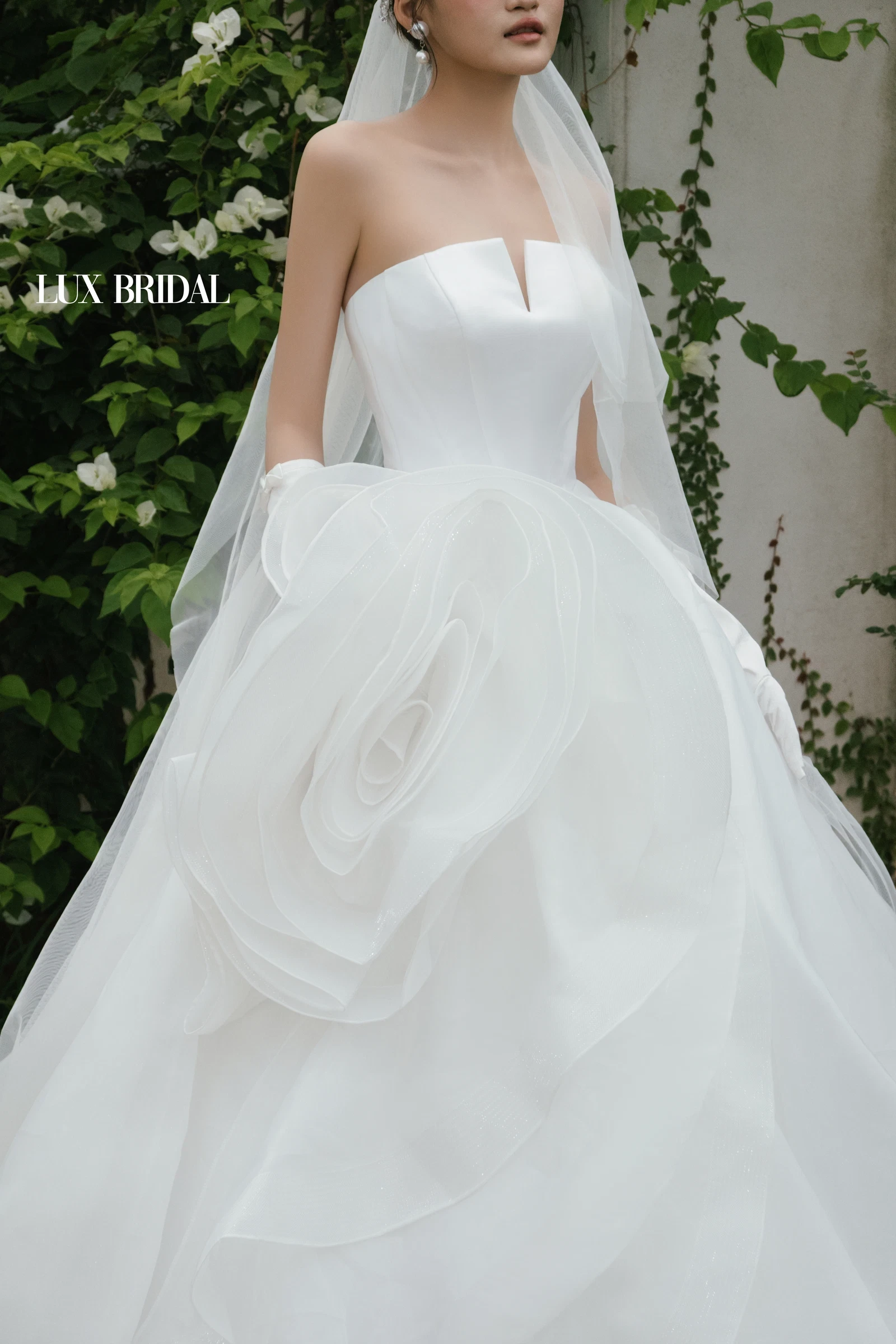 Cô dâu váy cưới trắng phần thân trơn, chân váy kết bông hoa organza lớn, đội voan dài đứng trước vườn cây xanh