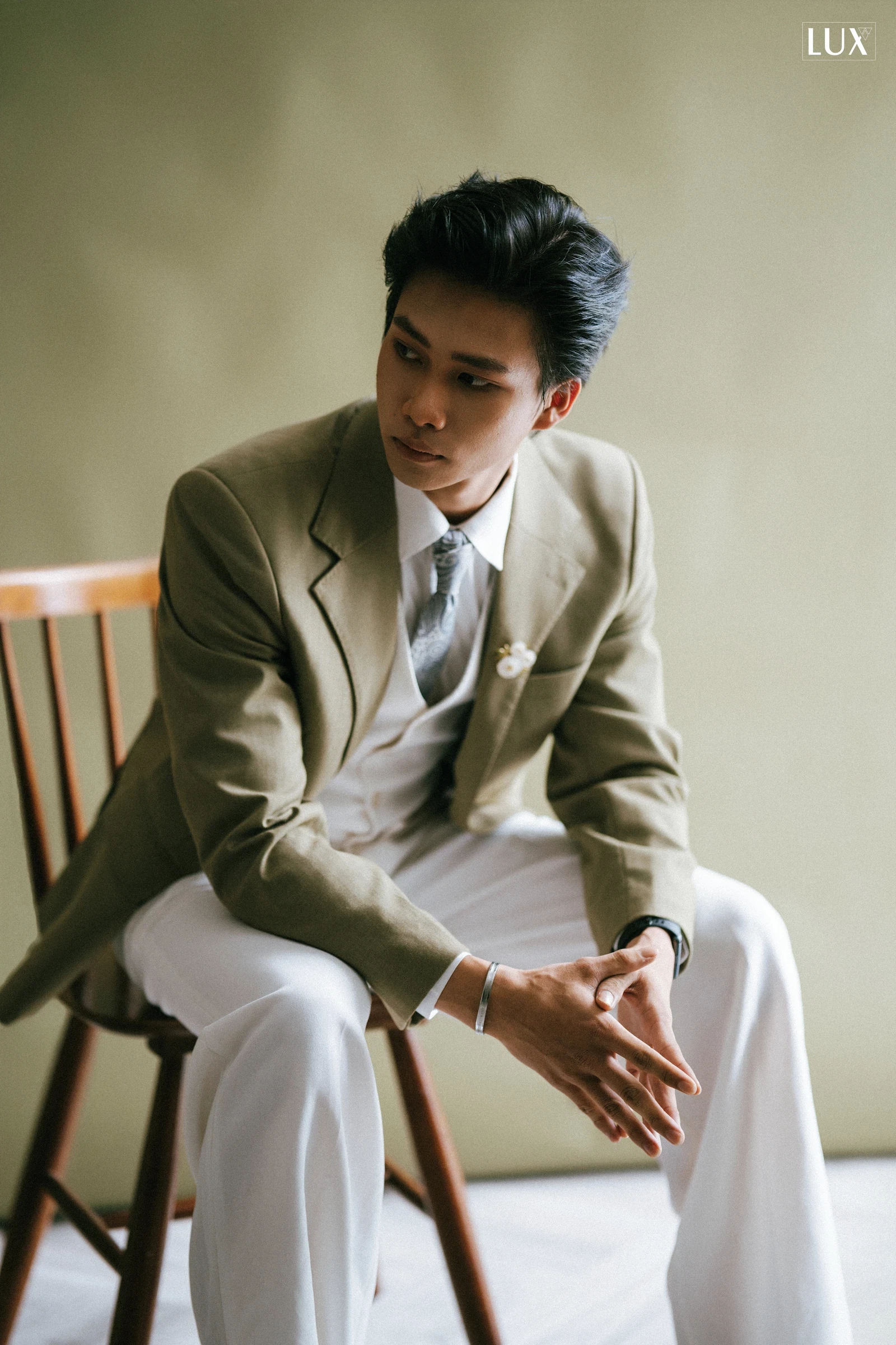 Chàng trai mặc blazer xanh rêu, sơ mi trắng và quần trắng ngồi trên ghế gỗ, hai tay đan vào nhau, ánh mắt nhìn xa, phông nền xanh rêu