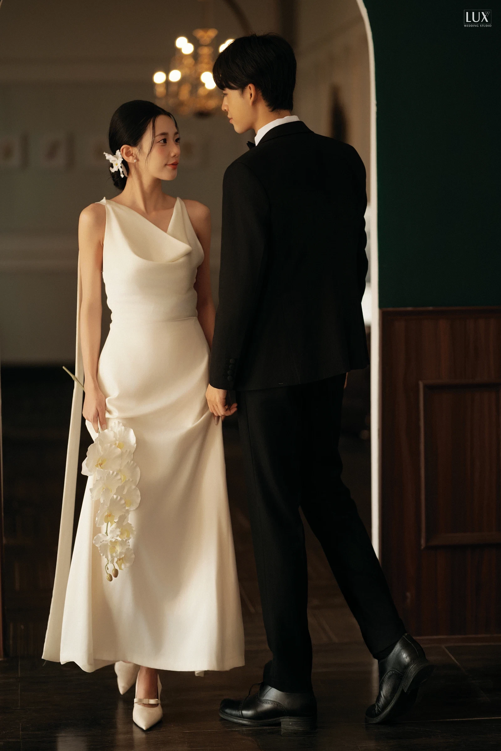Cô dâu váy satin trắng cổ đổ, cài hoa trên tóc, cầm cành lan trắng đứng nhìn chú rể vest đen dưới khung cửa vòm, tường xanh rêu và đèn chùm phía sau