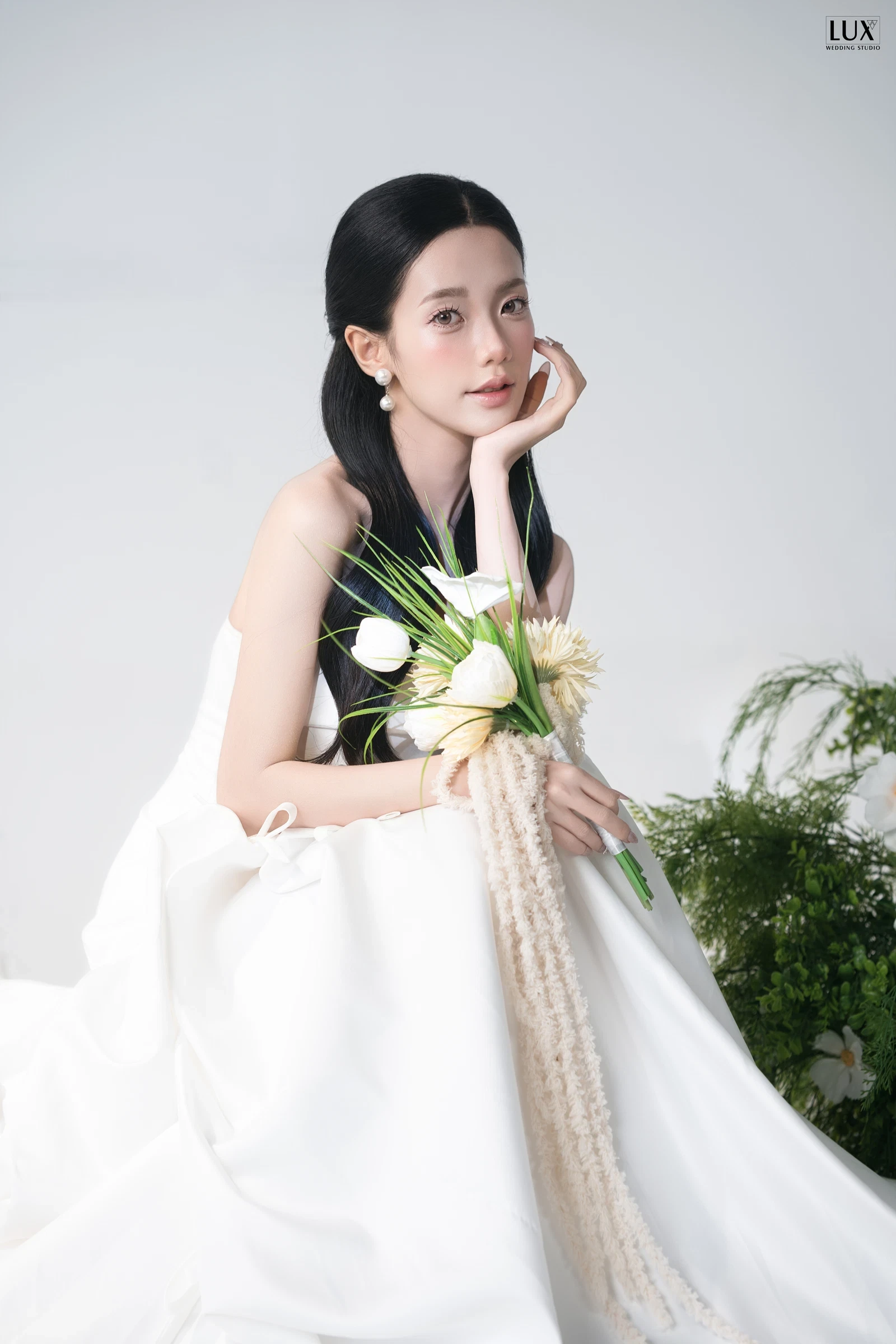 Cô dâu váy cưới trắng cúp ngực trơn ngồi tựa tay lên má, ôm bó hoa rum trắng, phông trắng bên khóm dương xỉ