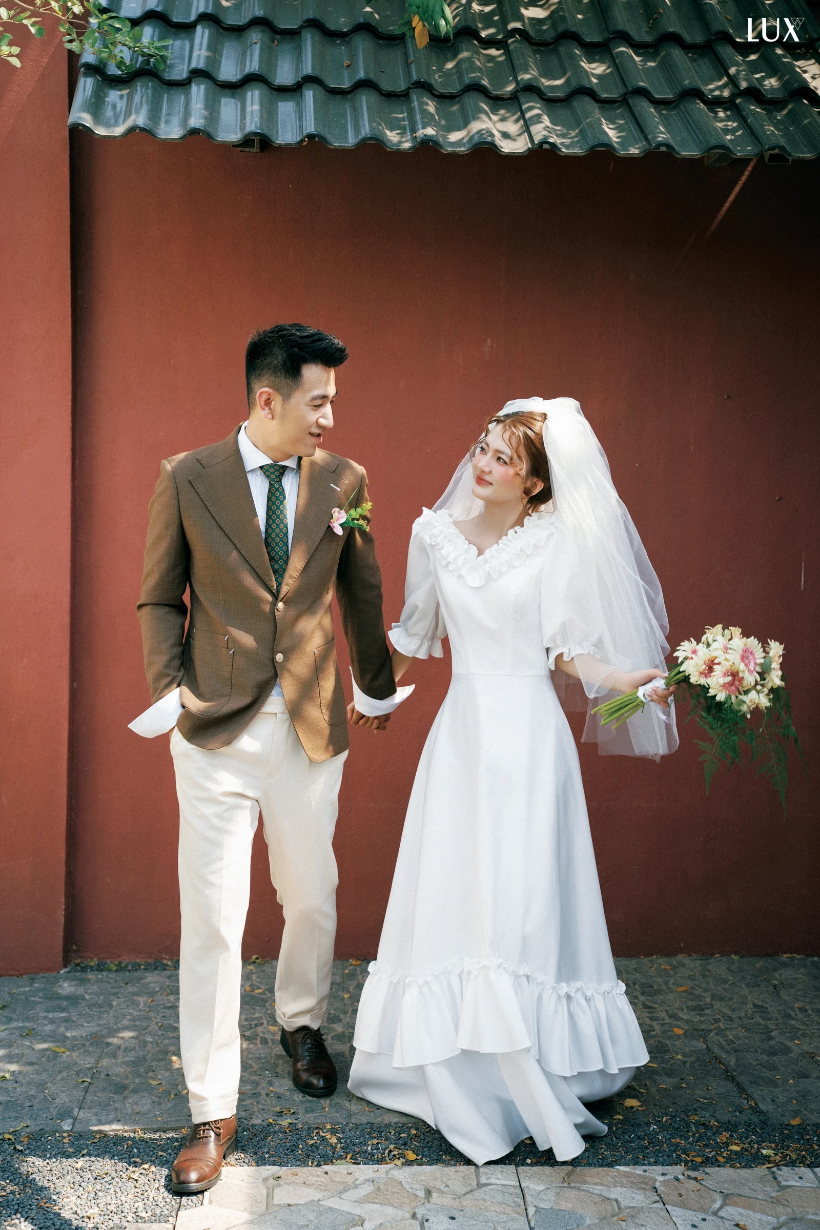 Chú rể áo vest nâu, quần trắng nắm tay cô dâu váy trắng đội voan, cùng cười rạng rỡ khi bước đi trước bức tường đỏ dưới mái ngói xanh