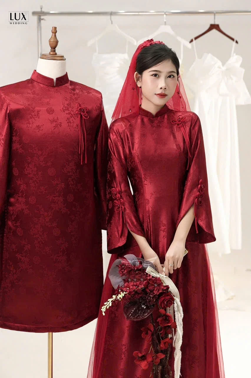 Cô dâu mặc áo dài cưới đỏ, đội khăn voan đỏ, cầm hoa đỏ, đứng cạnh chiếc áo dài nam đỏ trên ma-nơ-canh và giá treo váy trắng