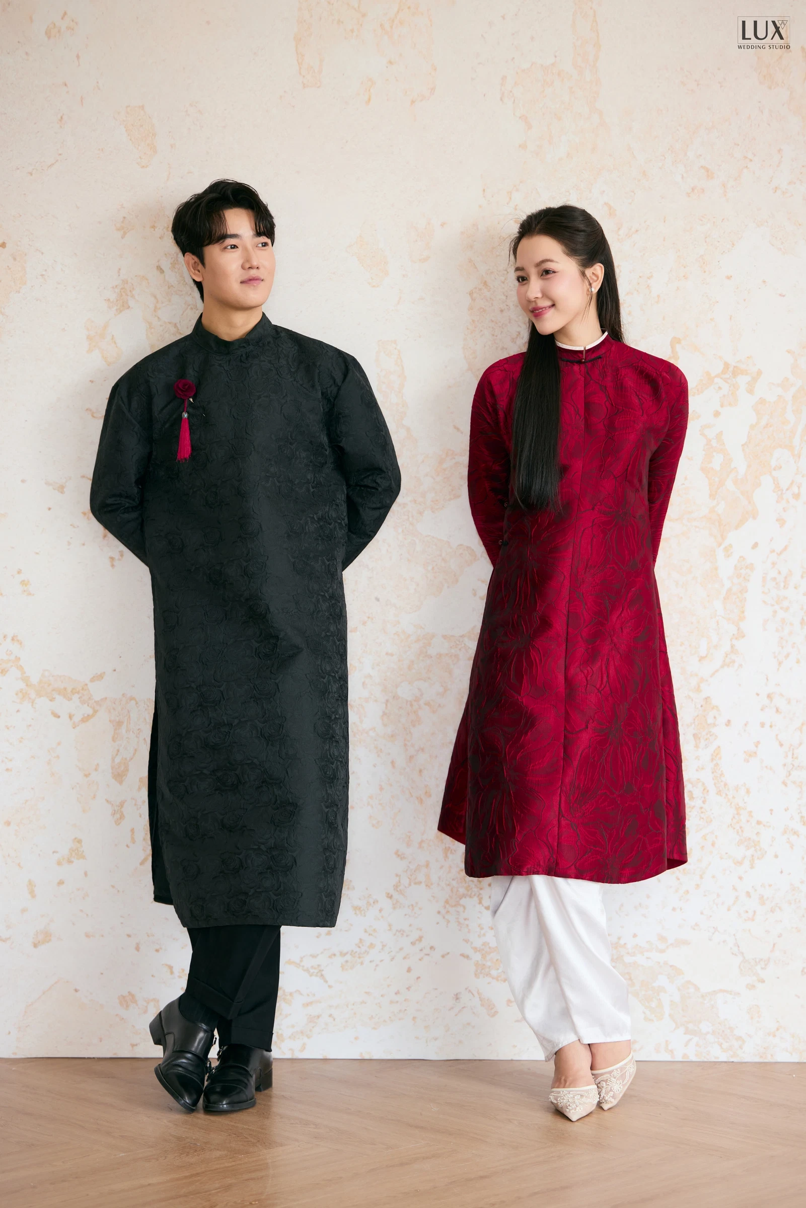 Chú rể áo dài đen hoa văn chìm, cài hoa và tua rua đỏ, đứng cạnh cô dâu áo dài đỏ đô, cả hai chắp tay sau lưng nhìn nhau trước phông màu kem