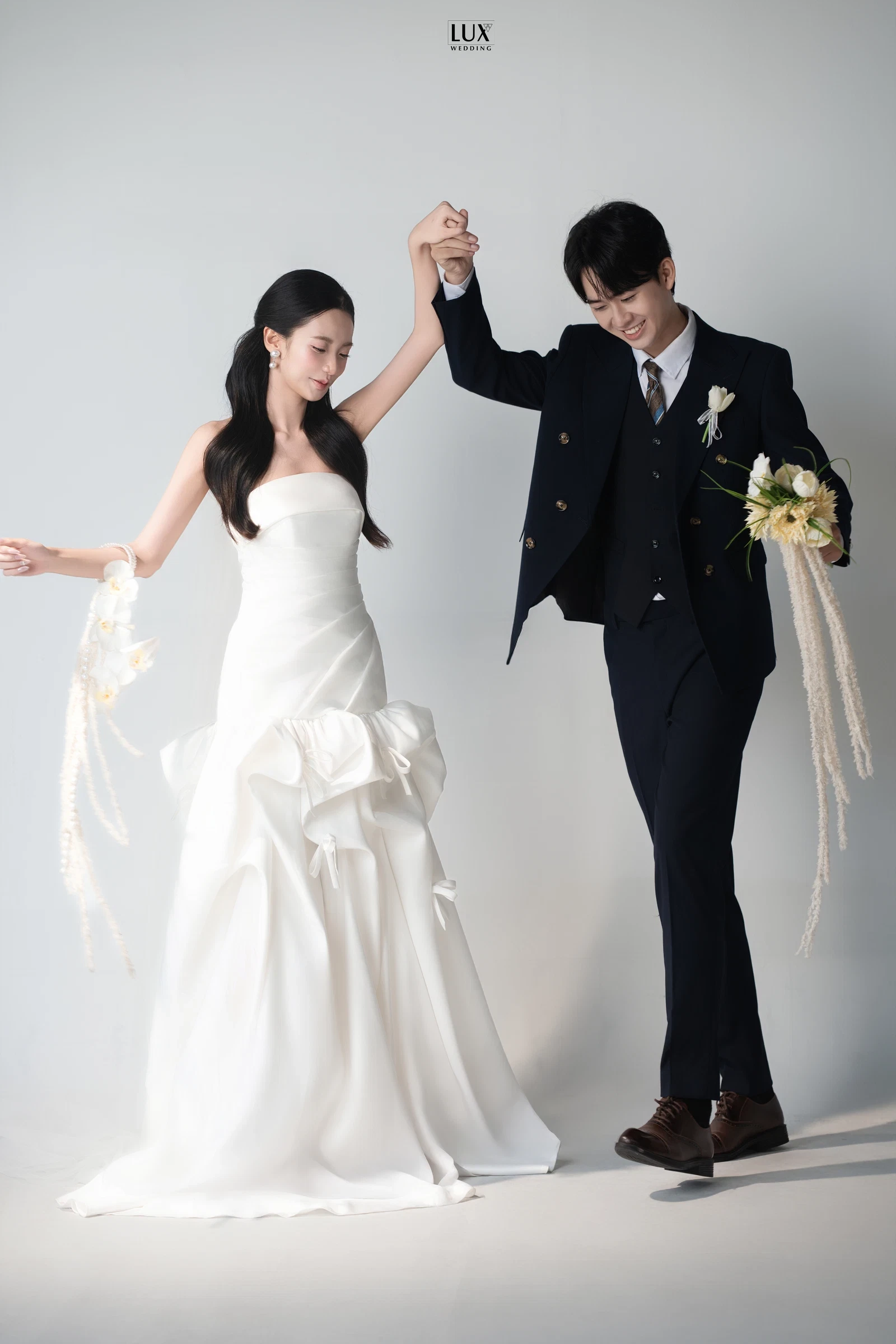 Cô dâu váy trắng cúp ngực đuôi cá, chú rể vest đen ba mảnh nắm tay nhau giơ cao, tươi cười trước phông trắng