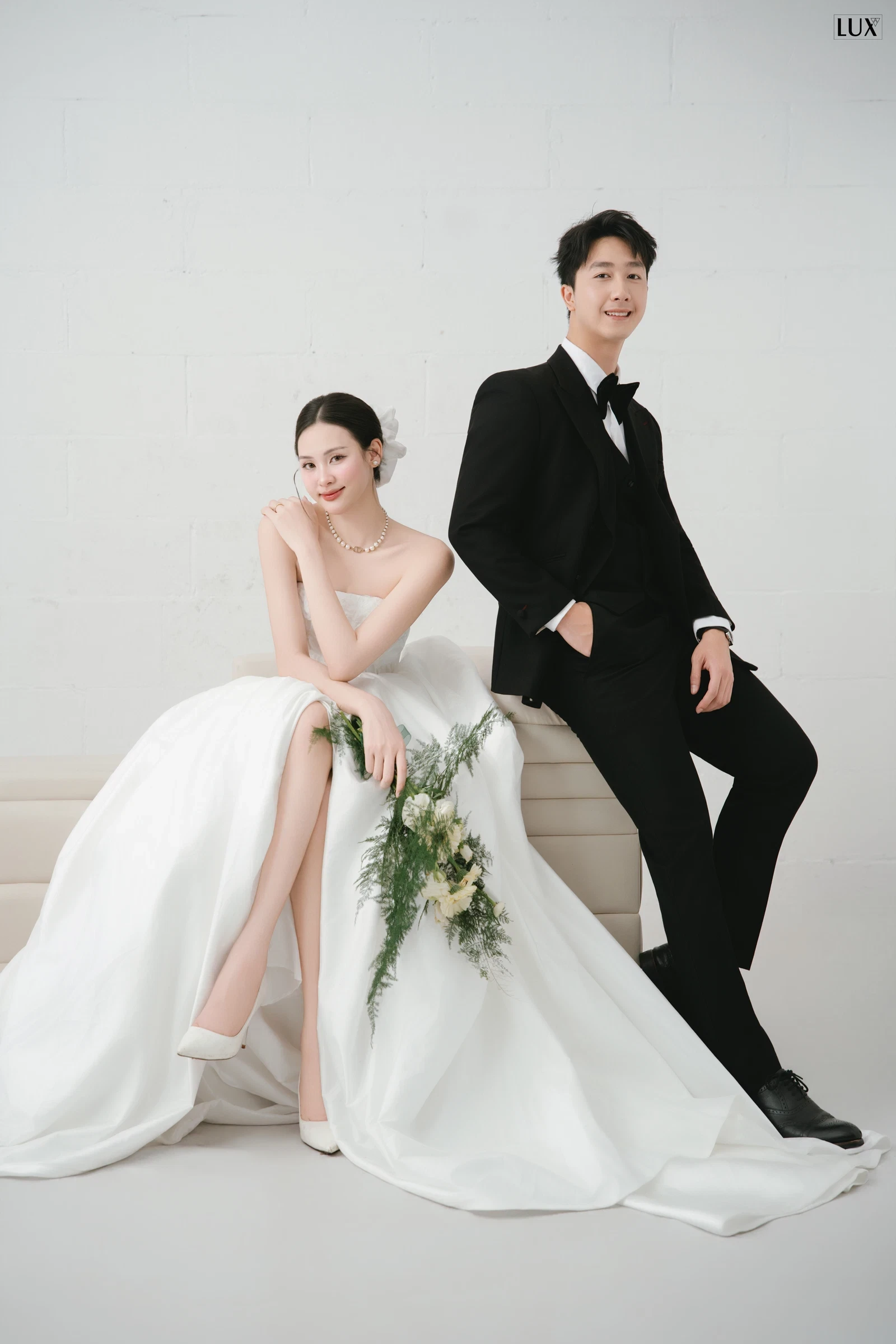 Cô dâu váy cúp ngực ngồi trên ghế dài màu kem, tay chống cằm, cầm bó hoa lá xanh; chú rể vest đen nơ đen ngồi tựa phía sau, một tay đút túi quần, cả hai mỉm cười nhìn về phía máy