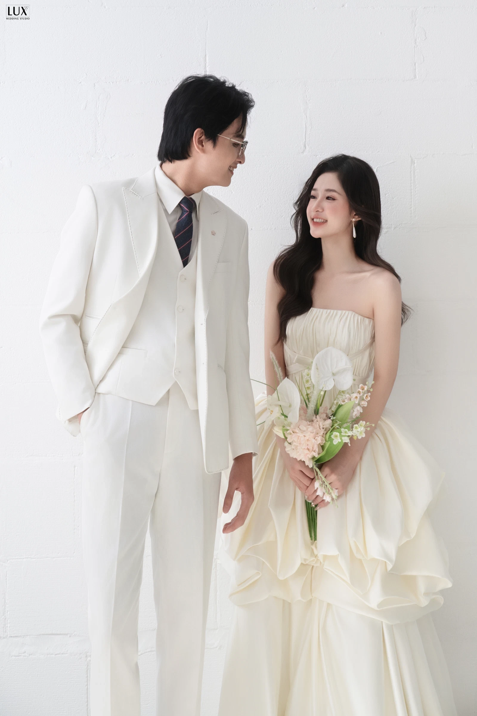 Cô dâu tóc xõa gợn sóng, trang điểm nhẹ nhàng, cầm bó hoa rum trắng, mỉm cười bên chú rể mặc vest trắng