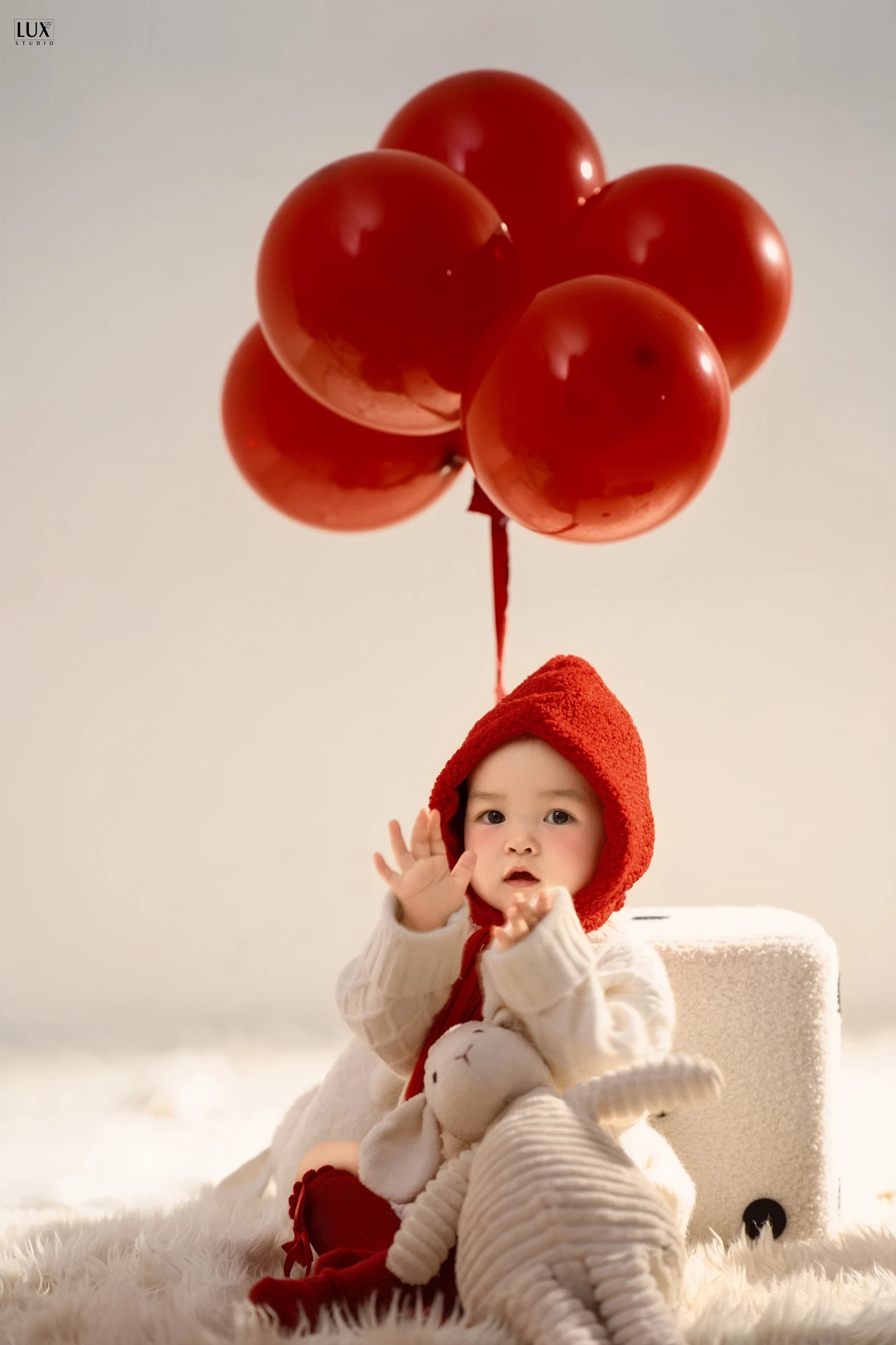 Bé đội mũ len đỏ ngồi trên thảm lông trắng, giơ tay vẫy, phía trên là chùm bóng bay đỏ, bên cạnh là thú bông