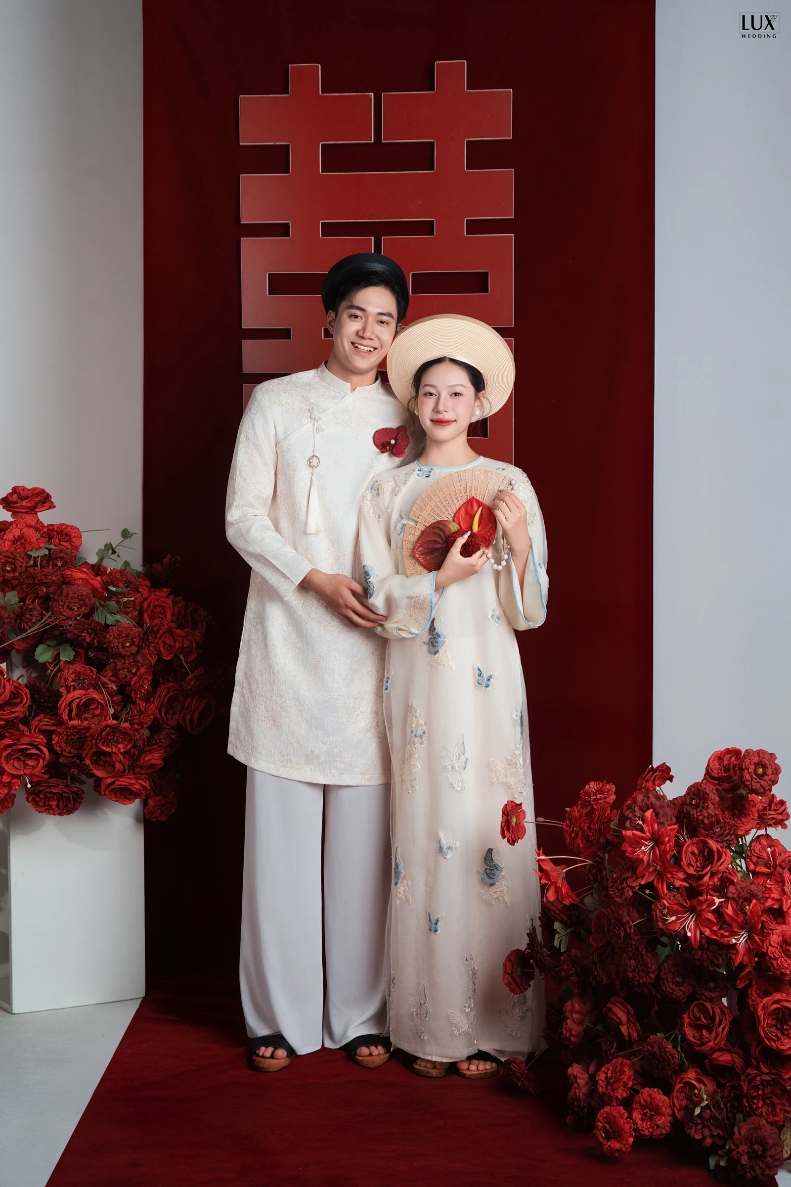Chú rể áo dài màu kem đội khăn xếp đen, ôm vai cô dâu mặc áo dài kem thêu bướm, đội nón tròn trắng, cầm quạt, bên cạnh chữ Hỷ đỏ và hoa hồng đỏ