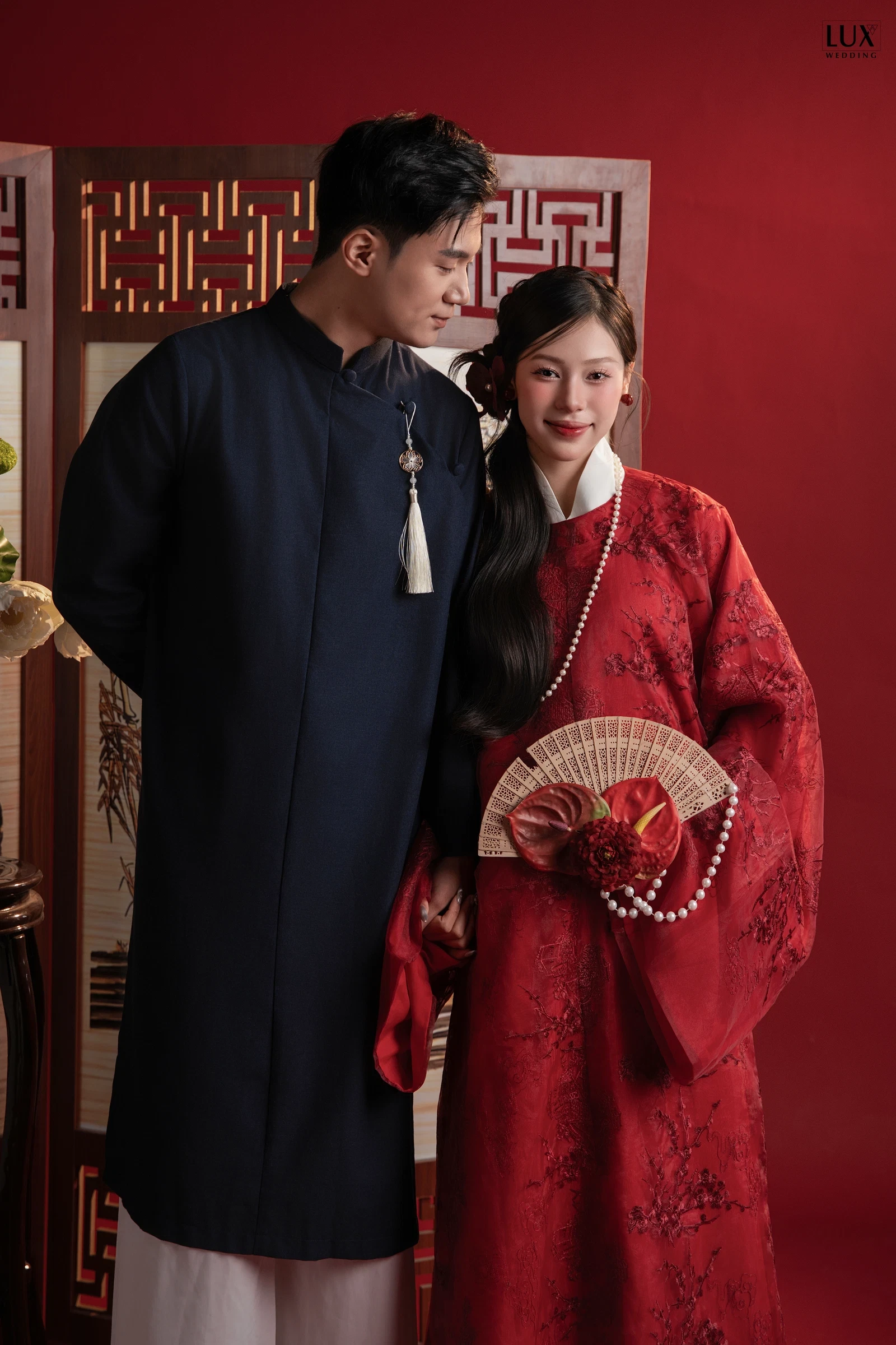 Cô dâu áo đỏ thêu hoa cầm quạt và chú rể áo dài xanh than đứng cạnh bình phong gỗ trước phông đỏ